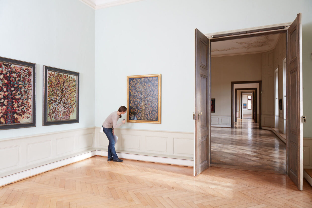 Sabine_Hofmeister_Ausstellungsbetreuung 1 - Deutsche Manufakturenstrasse Person überprüft ein Gemälde in einer Galerie, umgeben von weiteren Kunstwerken und einem hellen, offenen Raum.