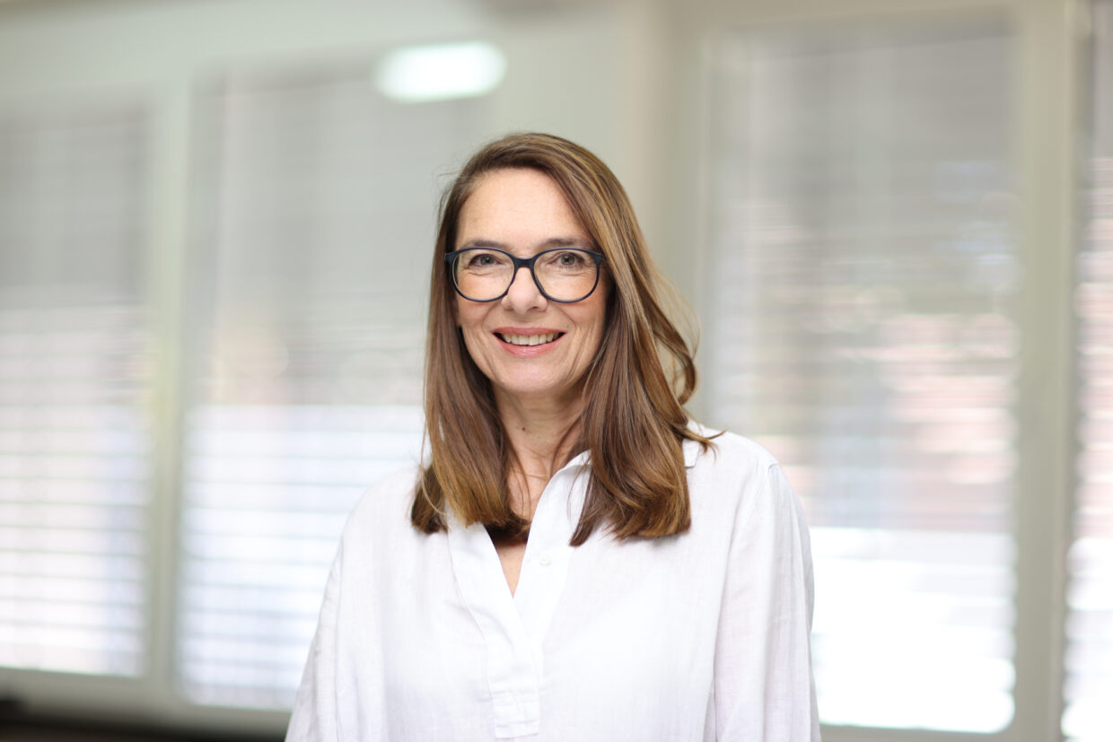 Sabine_Hofmeister_Portrait - Deutsche Manufakturenstrasse Porträt von Sabine Hofmeister mit langen, braunen Haaren und Brille, lächelnd vor einem hellen Hintergrund.