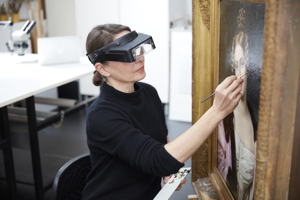 Sabine_Hofmeister_Retusche Leinwandgemälde - Deutsche Manufakturenstrasse Frau mit Vergrößerungsglas retuschiert ein Leinwandgemälde in einem Atelier.
