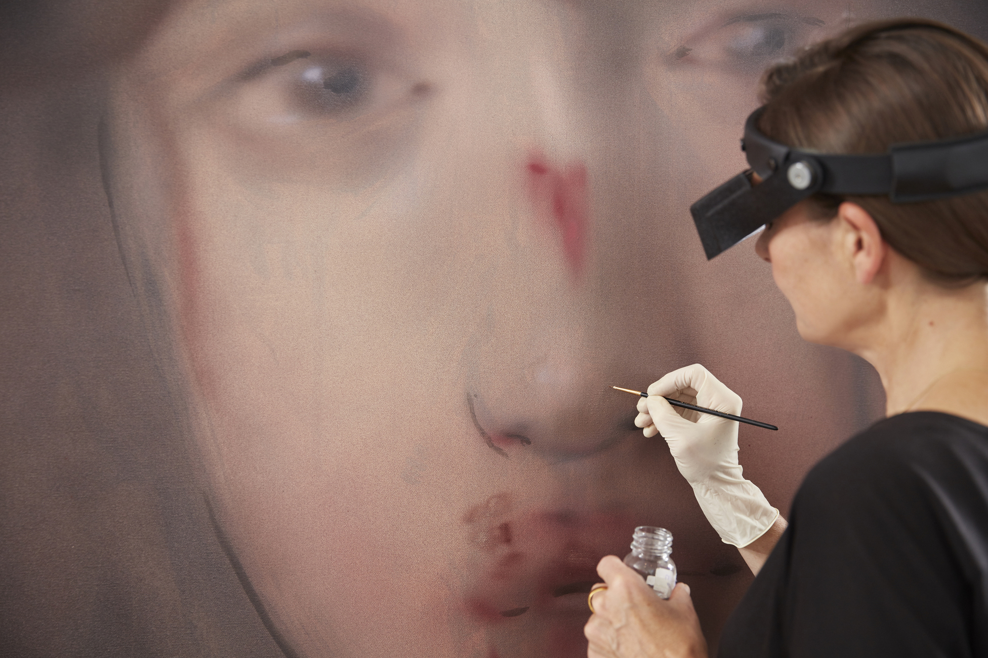 Sabine_Hofmeister_Retusche_Leinwandgemälde - Deutsche Manufakturenstrasse Künstlerin retuschiert ein Leinwandgemälde mit Fokus auf das Gesicht, trägt Handschuhe und eine Lupe.