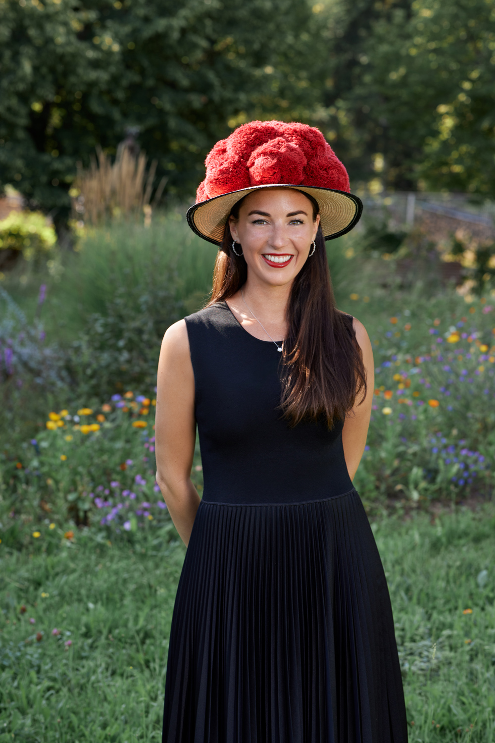 WEBAlicia-Lindner_BOERlIND©Sven-Cichowicz_02-2026_0I9A8523 - Deutsche Manufakturenstrasse Frau mit langen, dunklen Haaren in schwarzem Kleid und rotem, blühendem Hut steht vor einer bunten Blumenwiese.