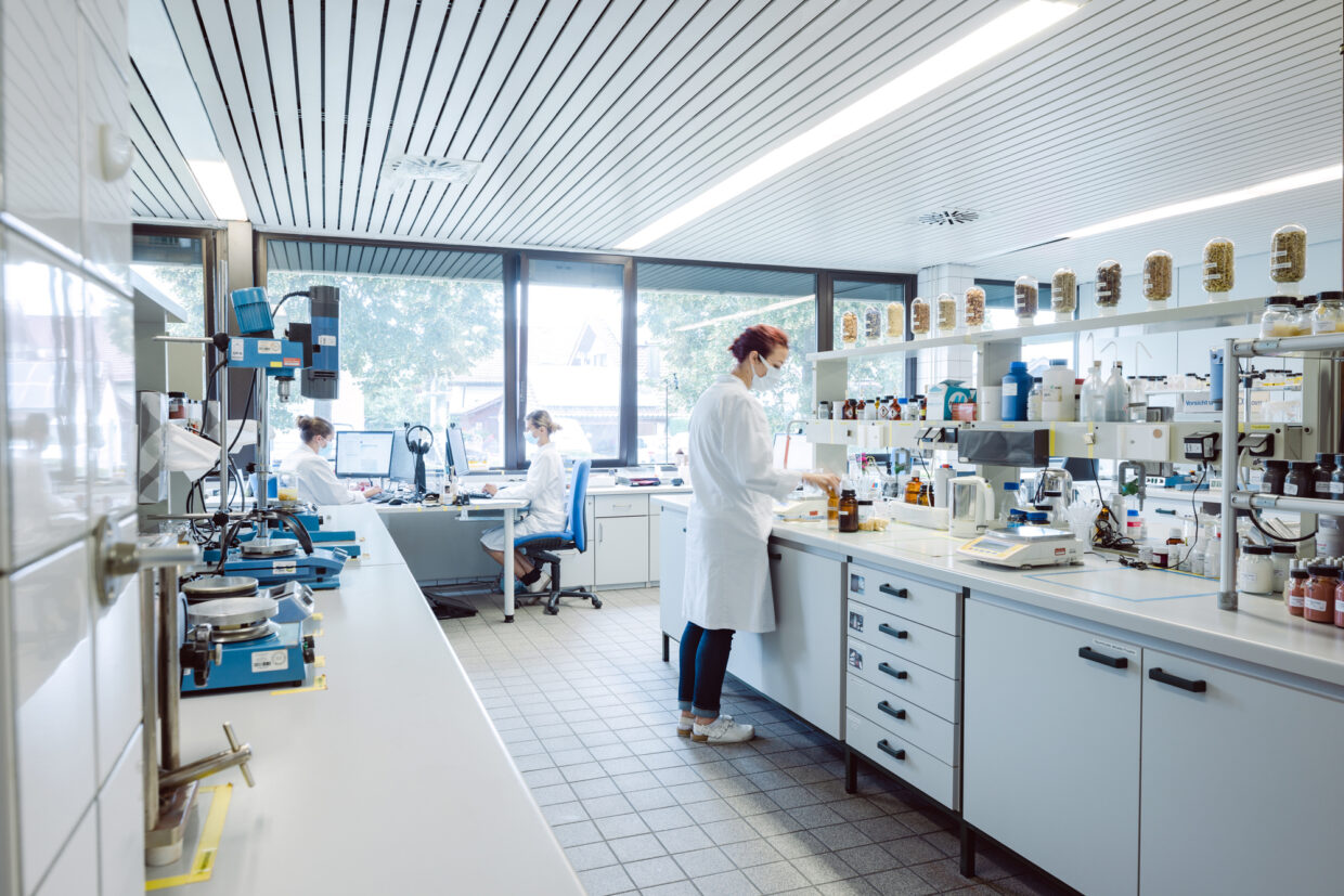 WEB_11A7171_Annemarie-Boerlind_Location_Sven-Cichowicz_2021 - Deutsche Manufakturenstrasse Laborumgebung mit drei Personen in weißen Laborkitteln, die an verschiedenen Arbeitsplätzen mit Geräten und Chemikalien...