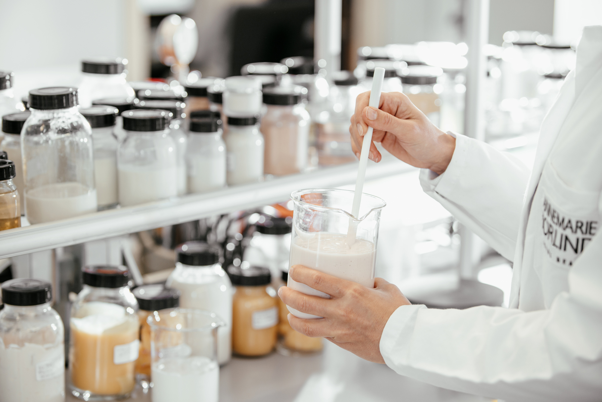 WEB_11A7245_Annemarie-Boerlind_Location_Sven-Cichowicz_2021 - Deutsche Manufakturenstrasse Laborumgebung mit einer Person in einem weißen Laborkittel, die eine Flüssigkeit in einem Becher rührt, umgeben von ver...
