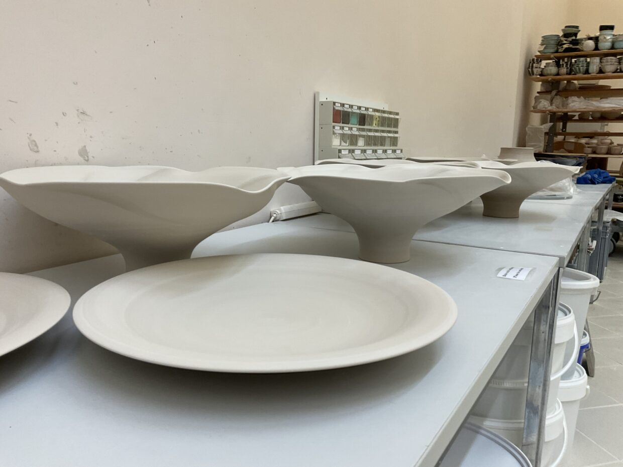 a2 1 - Deutsche Manufakturenstrasse Weißes Geschirr aus Keramik, darunter Schalen mit gewelltem Rand und flache Teller, auf einem Tisch in einem Atelier.