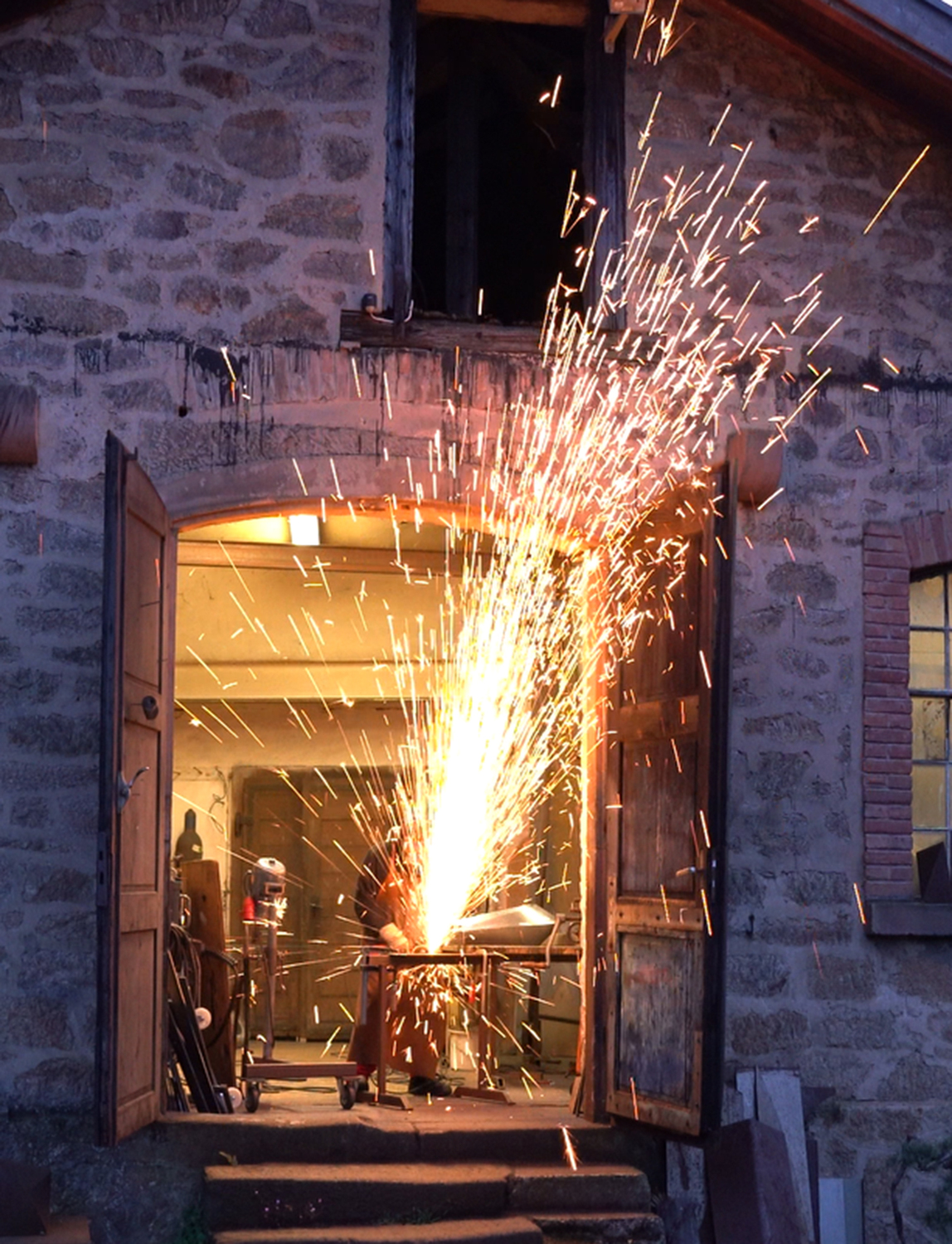 bei der Arbeit im Atelier 1 - Deutsche Manufakturenstrasse Funken sprühen aus einem Atelier, während eine Person an einem Metallobjekt arbeitet, umgeben von Steinmauern.