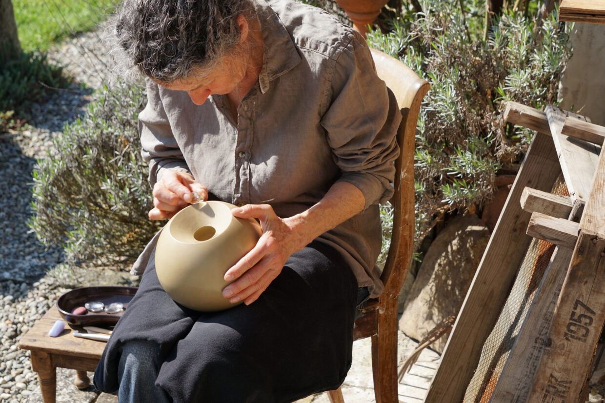 beim Polieren 1 - Deutsche Manufakturenstrasse Frau poliert einen beige Tontopf auf einem Stuhl im Freien, umgeben von Pflanzen und Holzpaletten.