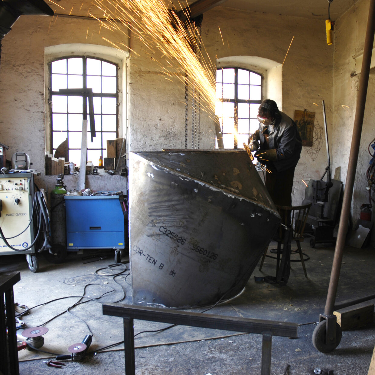 beim Schleifen 1 1 - Deutsche Manufakturenstrasse Handwerker schleift ein großes Metallobjekt in einer Werkstatt, Funken sprühen, Werkzeuge und Materialien sind im Hinter...