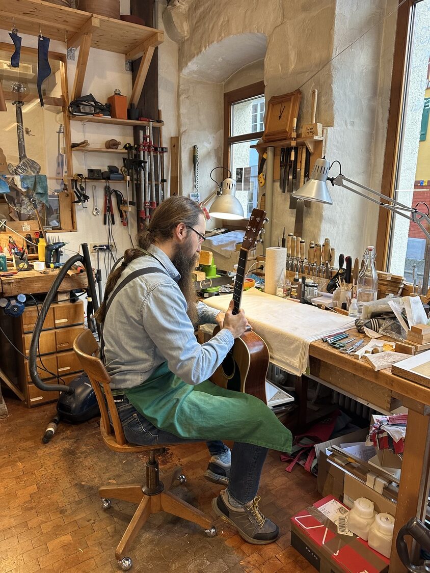 image00010 2 - Deutsche Manufakturenstrasse Gitarrenbauer sitzt in Werkstatt, arbeitet an einer Gitarre, umgeben von Werkzeugen und Materialien.
