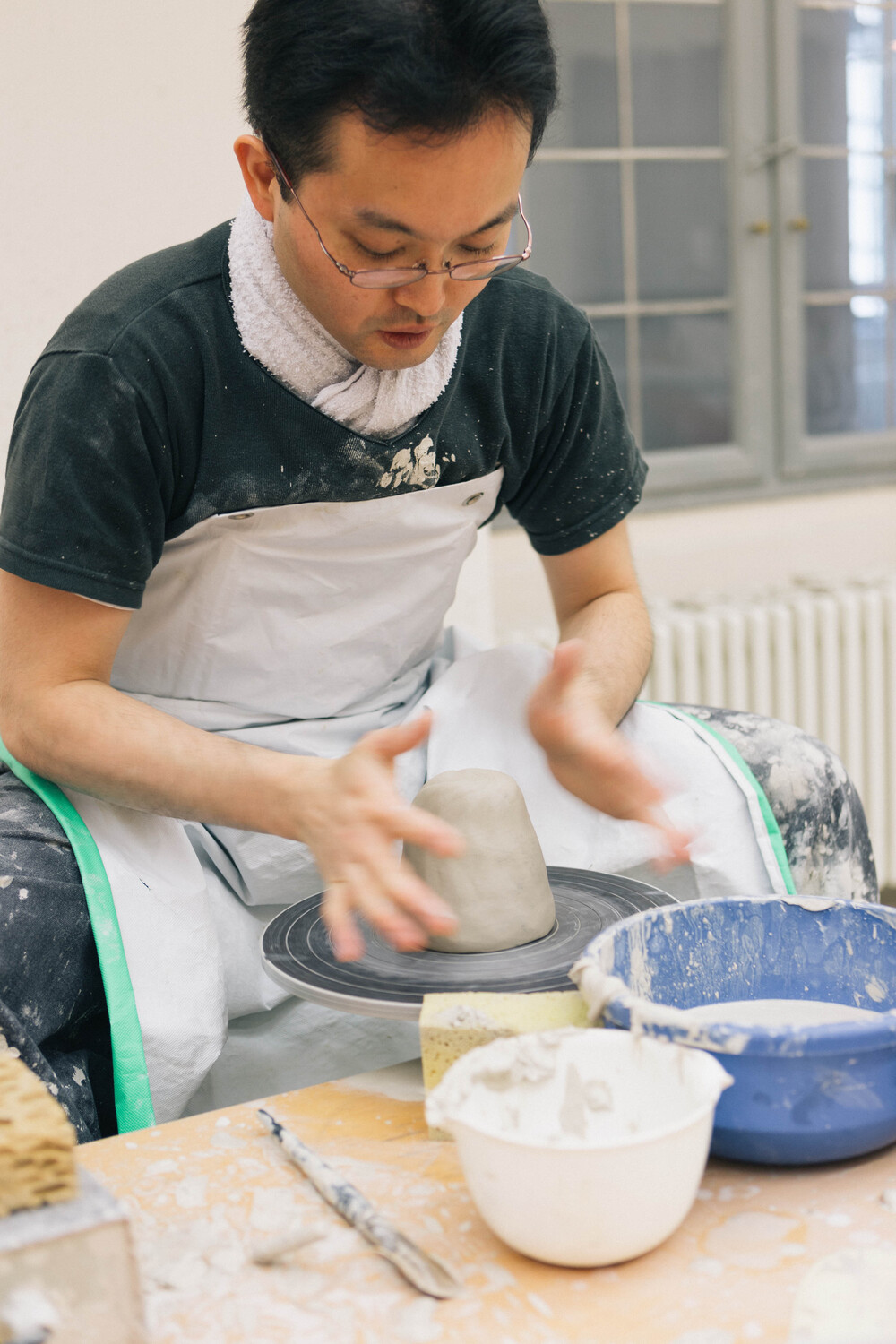 p1 1 - Deutsche Manufakturenstrasse Mann formt mit beiden Händen einen Tonklumpen auf einer Töpferscheibe, umgeben von Töpferwerkzeugen und Schalen.