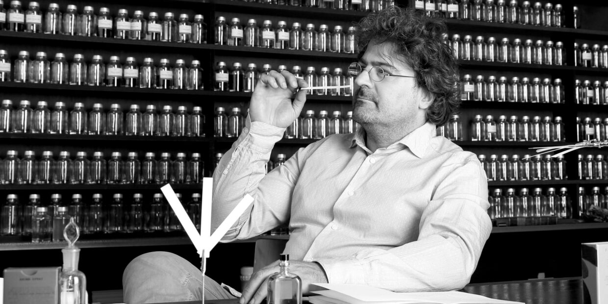 parfumeur portrait 1 - Deutsche Manufakturenstrasse Parfumeur sitzt an einem Tisch, riecht an einem Duftstoff, im Hintergrund eine Wand mit vielen Fläschchen.