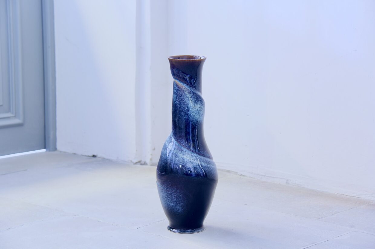 w2 1 - Deutsche Manufakturenstrasse Dunkelblaue, spiralförmige Vase aus Keramik steht auf einem hellen Boden vor einer Wand.