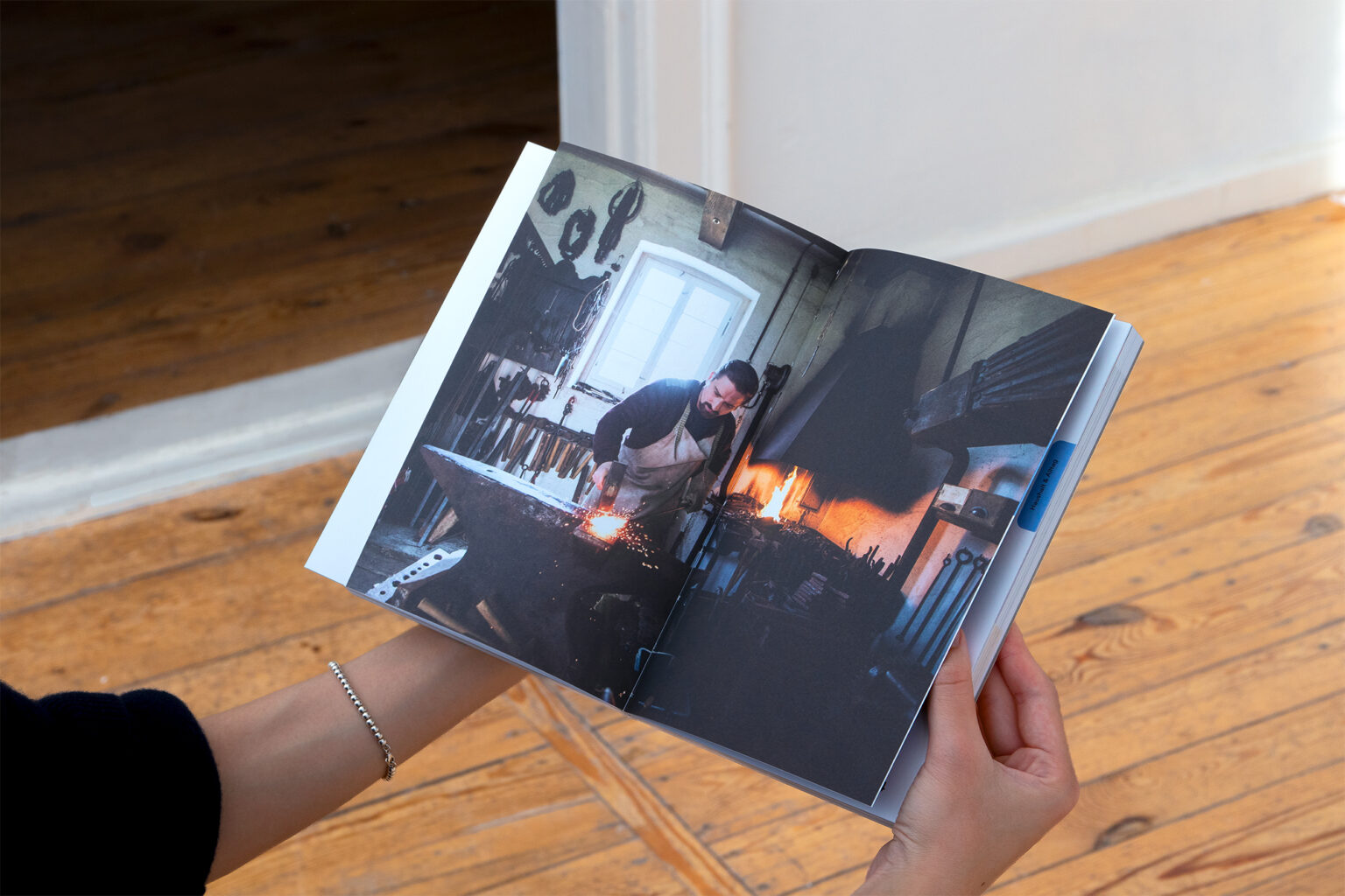 ManufaturenGuide_Teaser_web - Deutsche Manufakturenstrasse Hand hält ein aufgeschlagenes Buch, das einen Handwerker in einer Werkstatt zeigt, umgeben von Werkzeugen und einem Feuer.