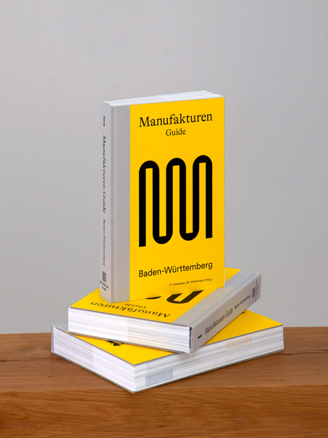 ManufaturenGuide_web_Variante_warm_shooting2-4-2_WEB - Deutsche Manufakturenstrasse Gelber Buchumschlag des "Manufakturen Guide Baden-Württemberg" auf einem Holzregal, gestapelt mit weiteren Exemplaren.